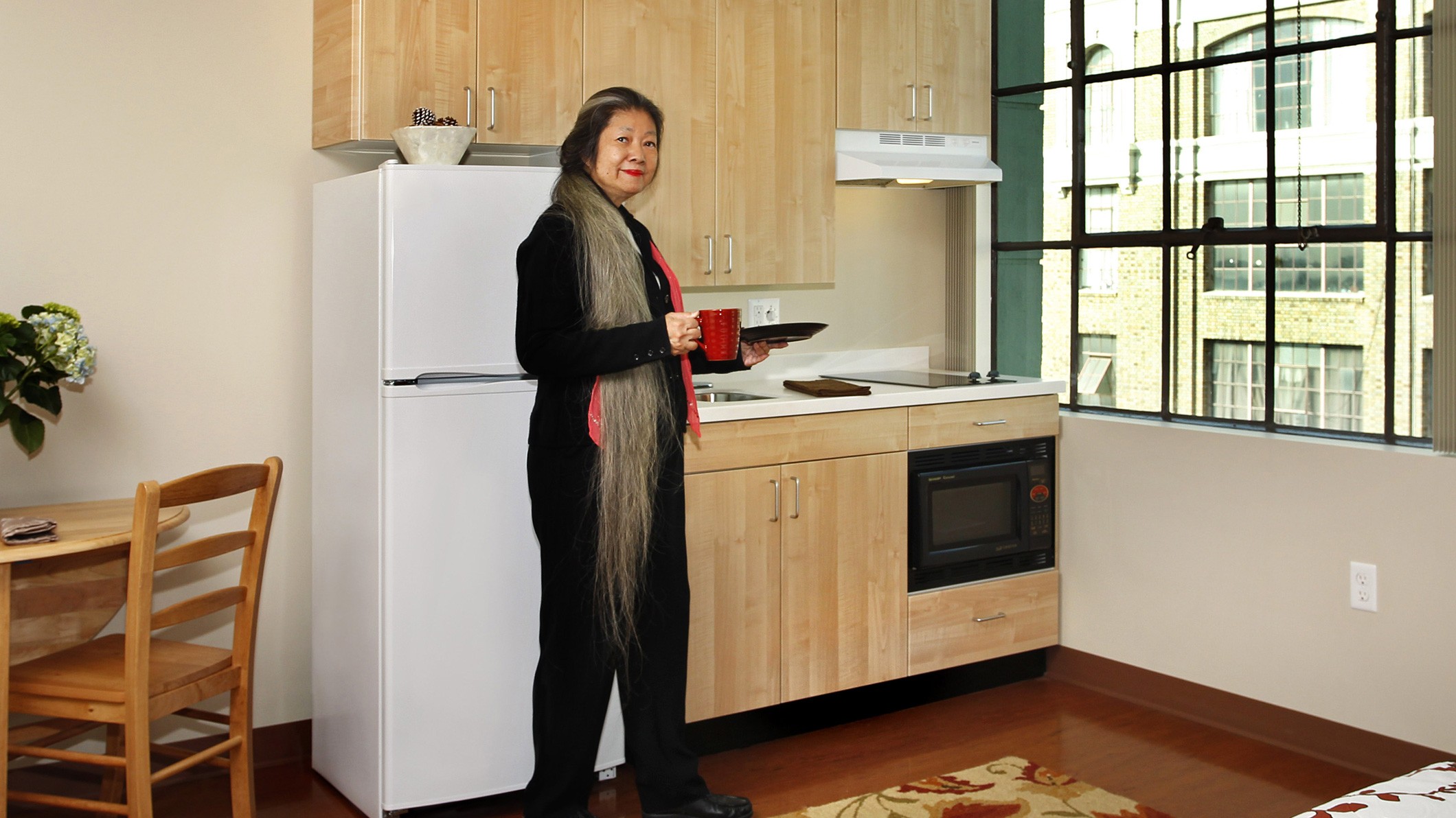 A woman stands with a plate and a mug in a kitchen at Downtown Women's Center in Los Angeles, a Pica+Sullivan Southern California non-profit architectural renovation project, from a low angle, focusing on the windows and decorative features.