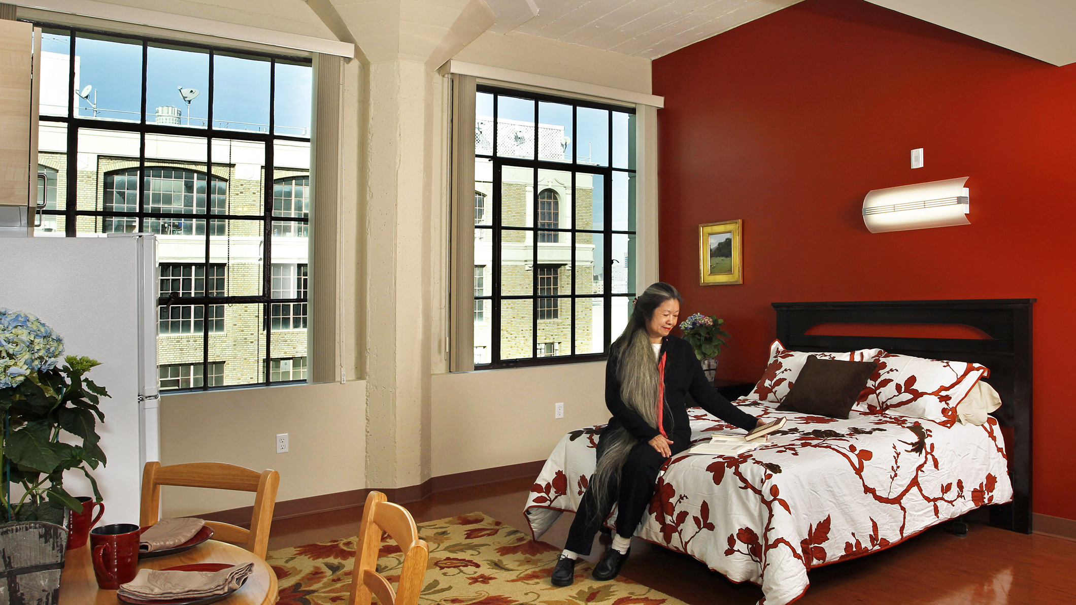 A woman sits on a bed in a comfortable room in the Downtown Women's Center in Los Angeles, a Pica+Sullivan Southern California non-profit architectural renovation project.
