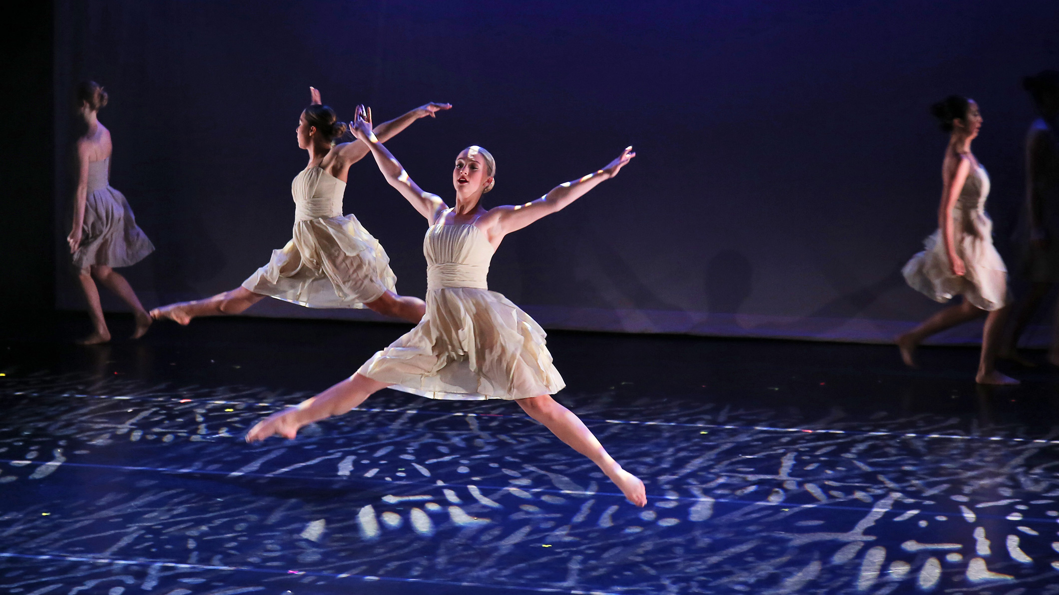 Two female student ballerinas leap into the air on stage at the Mozilo Family Center for the Arts at the Flintridge Sacred Arts Academy, an educational architectural project in Southern California.
