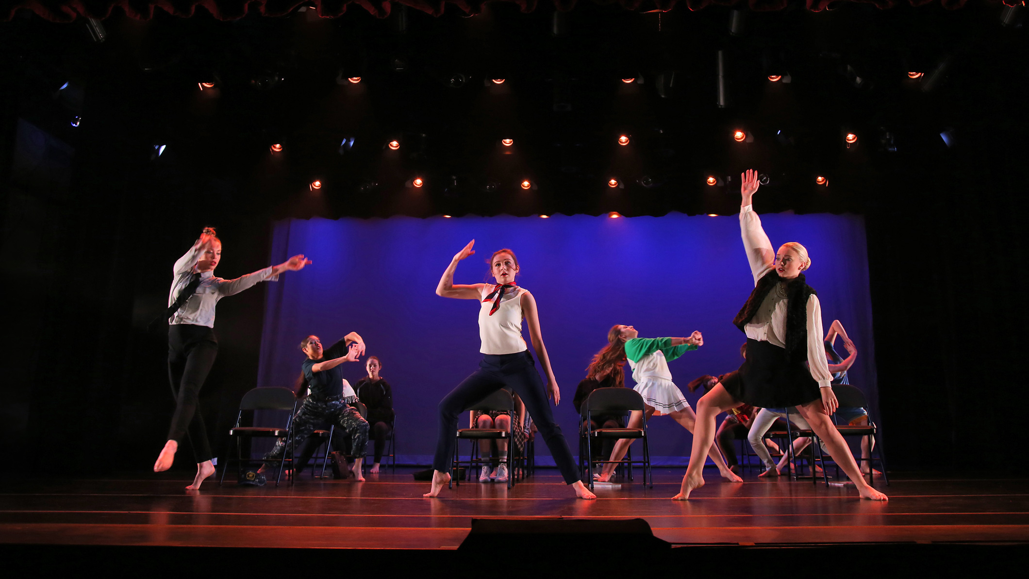 Female students dance on stage dressed in modern clothes at the Mozilo Family Center for the Arts at the Flintridge Sacred Arts Academy, an educational architectural project in Southern California. A number of folding chairs in rows of four are in the middle of the dancing students on stage.