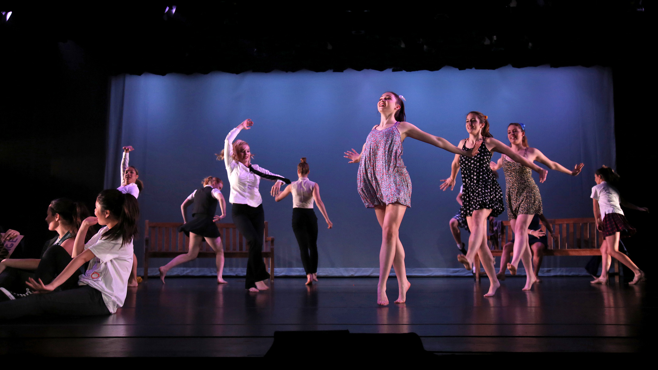 Female students dance on stage dressed in modern clothes at the Mozilo Family Center for the Arts at the Flintridge Sacred Arts Academy, an educational architectural project in Southern California. Two benches are on stage behind them on the left and right. A figure is seatedon the right bench.
