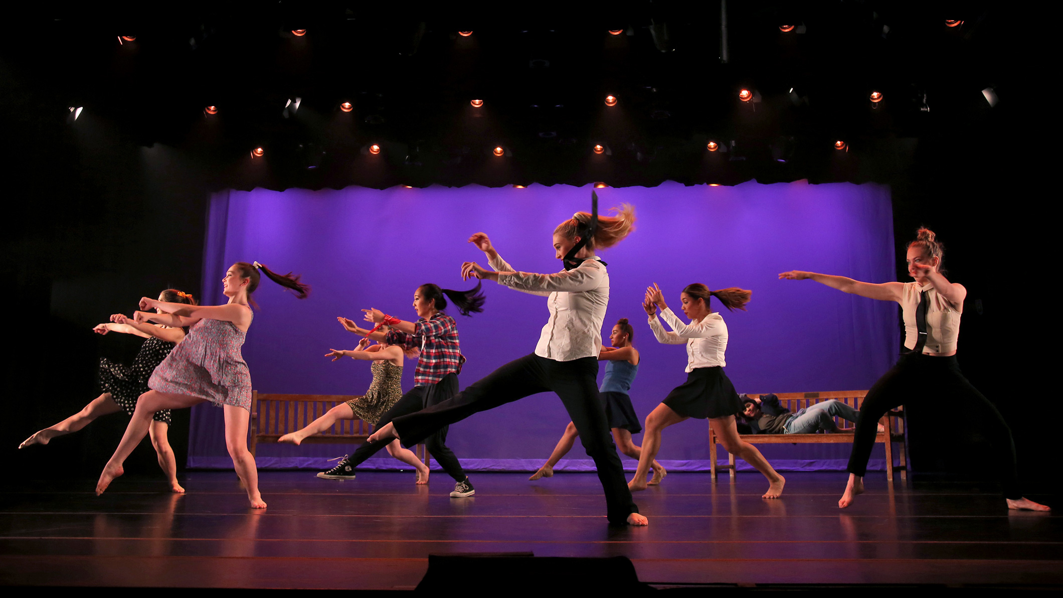 Female students dance on stage dressed in modern clothes at the Mozilo Family Center for the Arts at the Flintridge Sacred Arts Academy, an educational architectural project in Southern California. Two benches are on stage behind them on the left and right. A figure is lying on the right bench.
