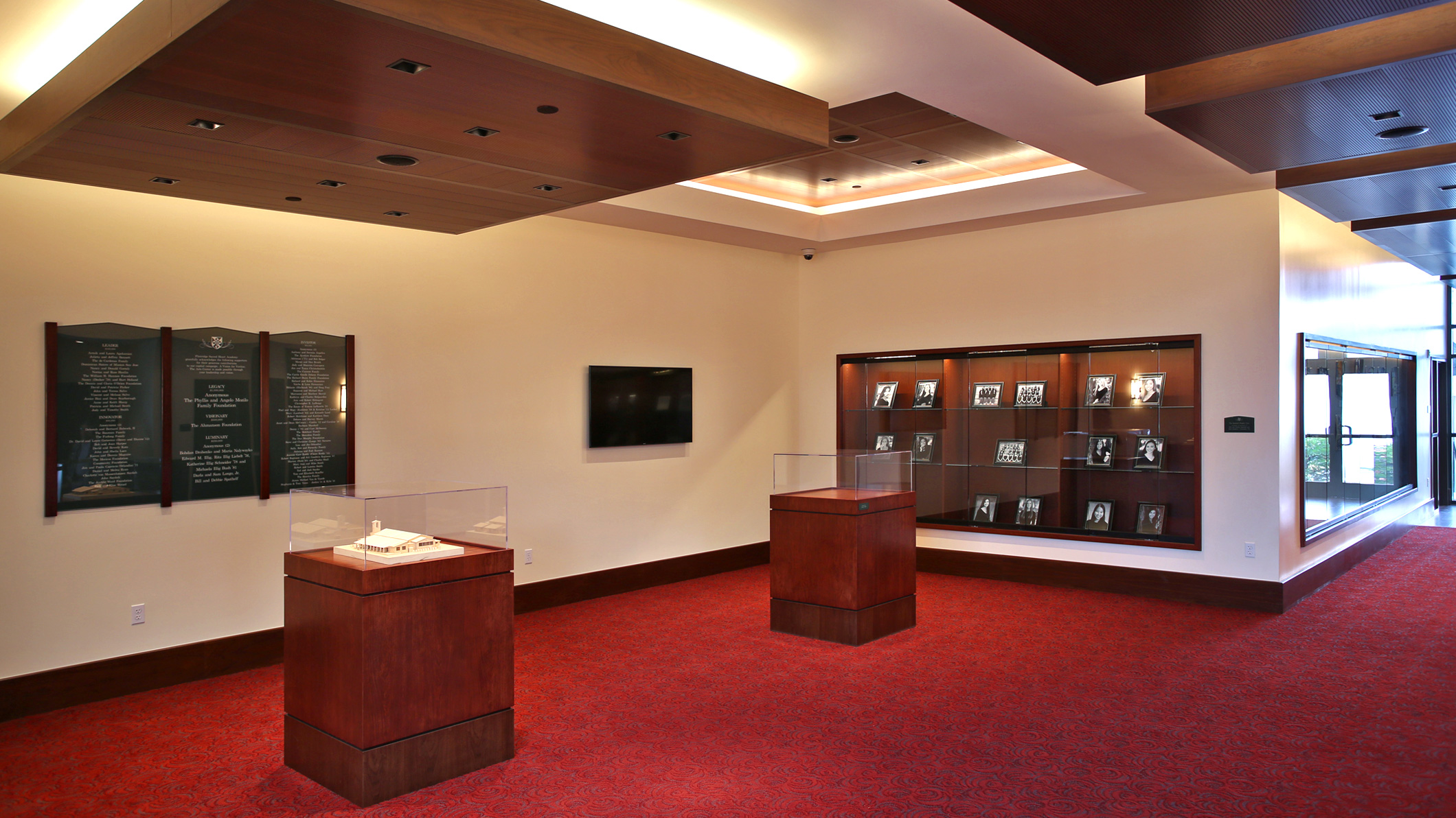 An interior display room at the Mozilo Family Center for the Arts at the Flintridge Sacred Arts Academy, an educational architectural project in Southern California. Two wooden display cases with glass tops sit in the center of the room, while a larger display case is recessed in the wall containing photographs. Signs and a TV are on the adjacent wall to the larger display case.
