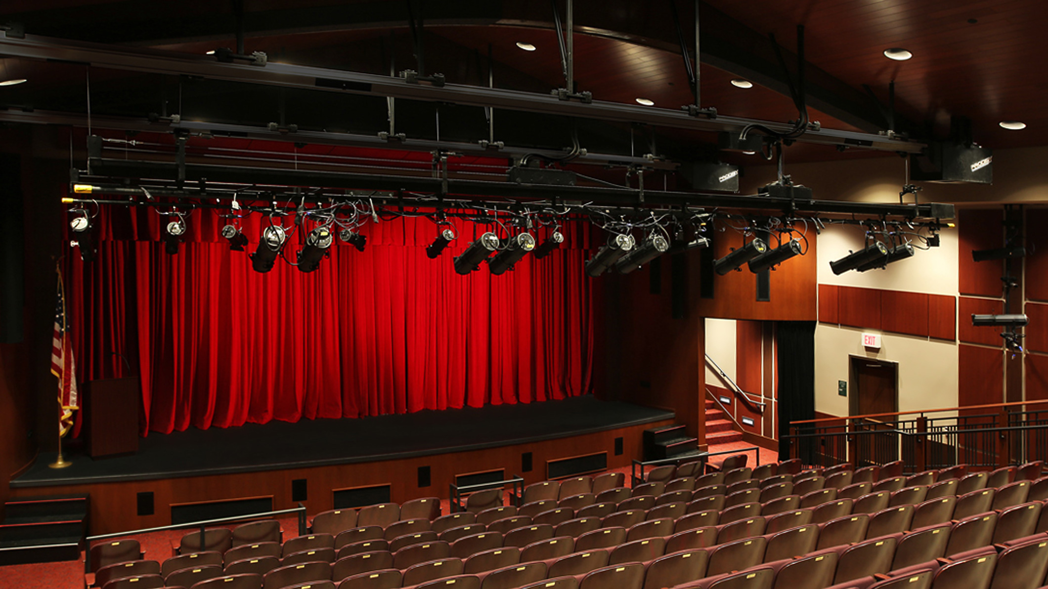 View of the stage from the top row, stage-left, of an auditorium at the Mozilo Family Center for the Arts at the Flintridge Sacred Arts Academy, an educational architectural project in Southern California. Theater lights are visible above, while the red curtain is closed on stage. A staircase to the right of the stage leads back.