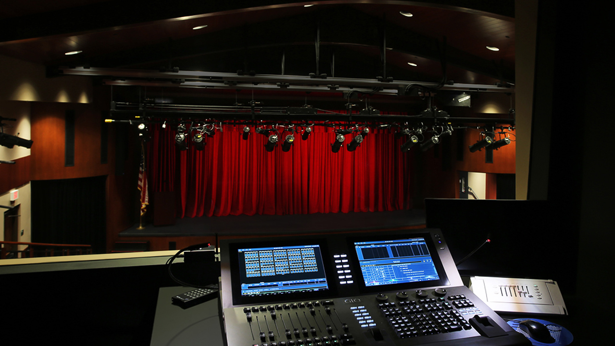 View of the stage from the tech booth inside an auditorium at the Mozilo Family Center for the Arts at the Flintridge Sacred Arts Academy, an educational architectural project in Southern California. A lighting board sits on a table in front of a window where the theater lights and closed red curtain are visible