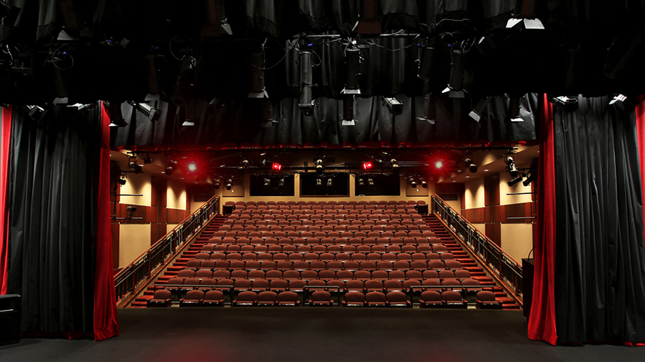 View of an auditorium from the stage inside the Mozilo Family Center for the Arts at the Flintridge Sacred Arts Academy, an educational architectural project in Southern California. Curtains frame the seats, black on the interior and red on the exterior. Stage lights above light the room.