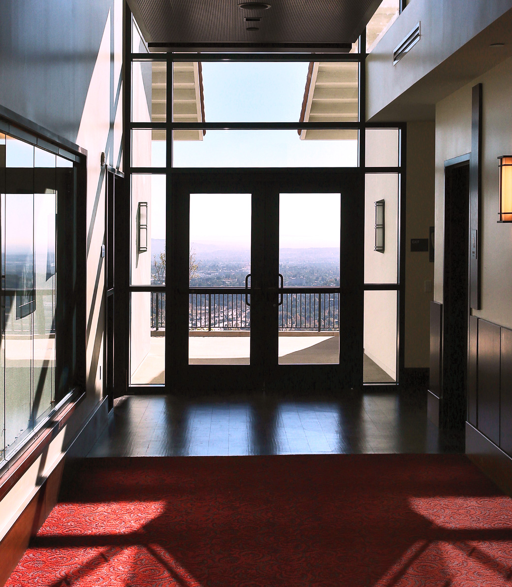 Expanded Mozilo Family Center for the Arts – Pica + Sullivan.com Interior view of a door leading out to a landing at the Mozilo Family Center for the Arts at the Flintridge Sacred Arts Academy, an educational architectural project in Southern California. A view of the surrounding neighborhood and forests is visible through the glass windows and doors.