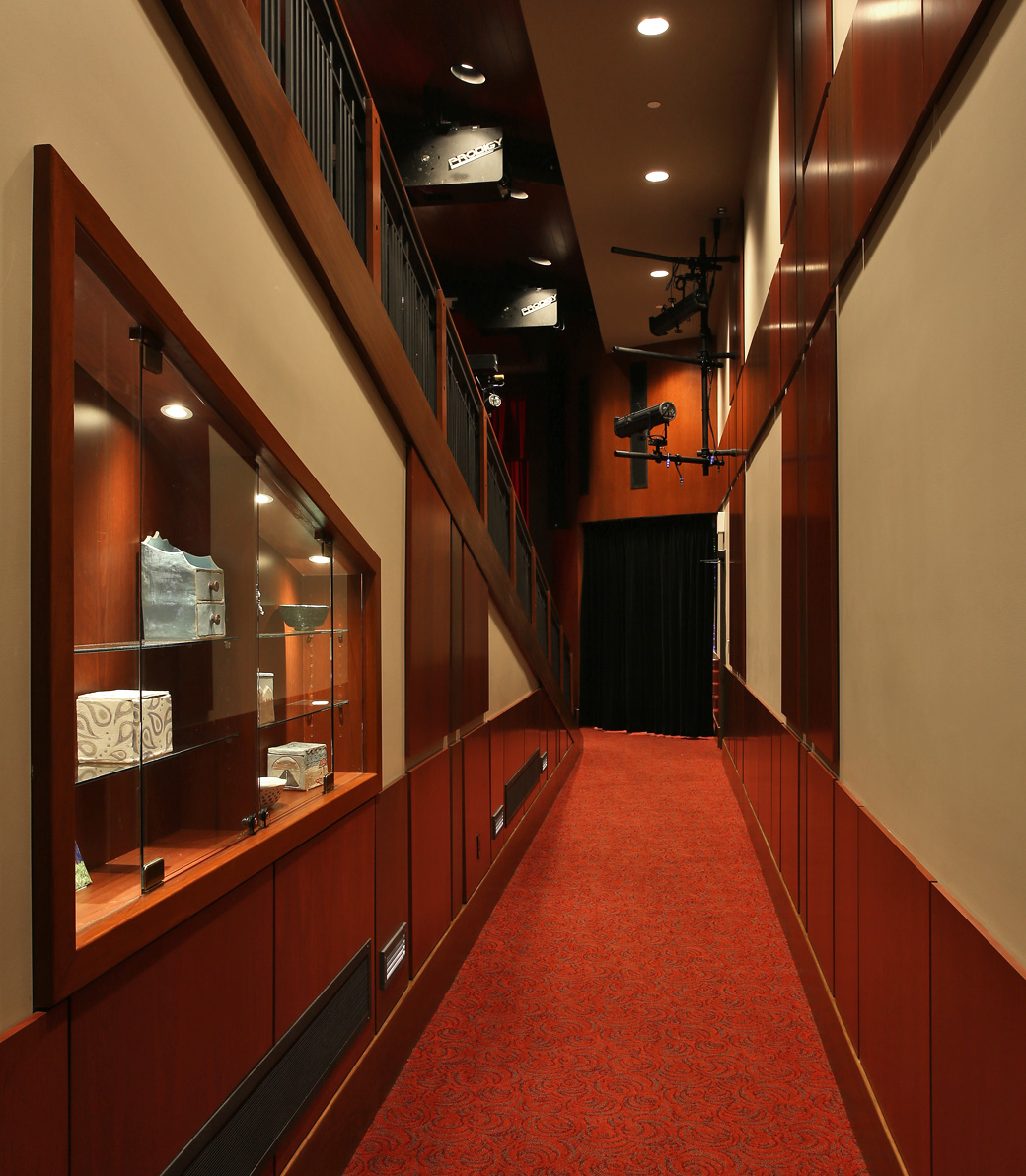 Expanded Mozilo Family Center for the Arts – Pica + Sullivan.com An interior hallway in the Mozilo Family Center for the Arts at the Flintridge Sacred Arts Academy, an educational architectural project in Southern California. Red floral-patterned carpet on the floor, a glass case with art items on the left, and stage-style lights above.