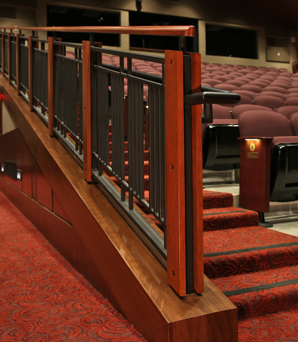Expanded Mozilo Family Center for the Arts – Pica + Sullivan.com Close-up of a handrail inside an auditorium at the Mozilo Family Center for the Arts at the Flintridge Sacred Arts Academy, an educational architectural project in Southern California. The handrail is dark metal bars with red wood accents. The auditorium chairs are visible on the right and a hallway leads off to the left. The carpet is a red floral pattern.