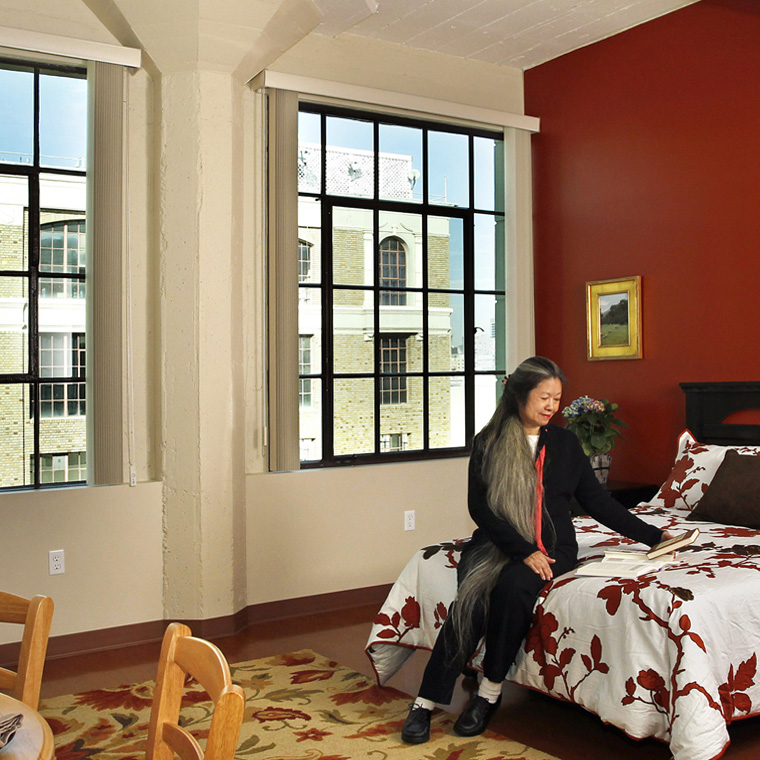 A woman sits on a bed in a comfortable room in the Downtown Women's Center in Los Angeles, a Pica+Sullivan Southern California non-profit architectural renovation project.