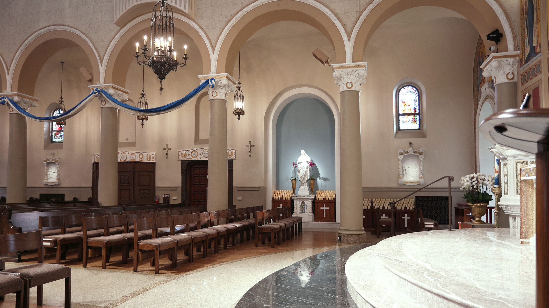 The empty pews of Saint Monica Church in Santa Monica, a Pica+Sullivan Southern California religious architectural renovation project, with a marble statue, devotional candles, and confessionals in the background.