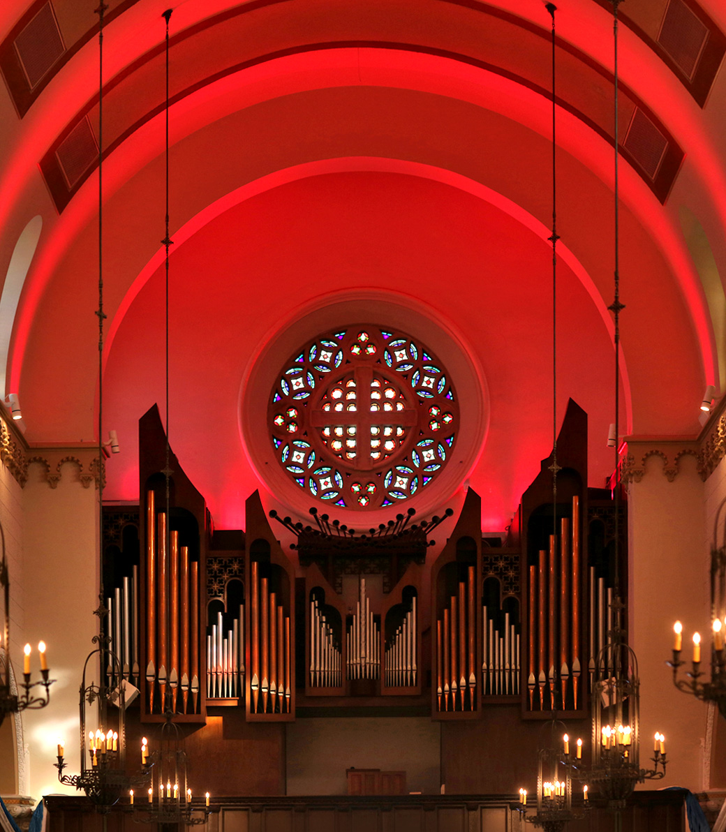 The pipe organ and stained glass window lit in red at Saint Monica Church in Santa Monica, a Pica+Sullivan Southern California religious architectural renovation project.