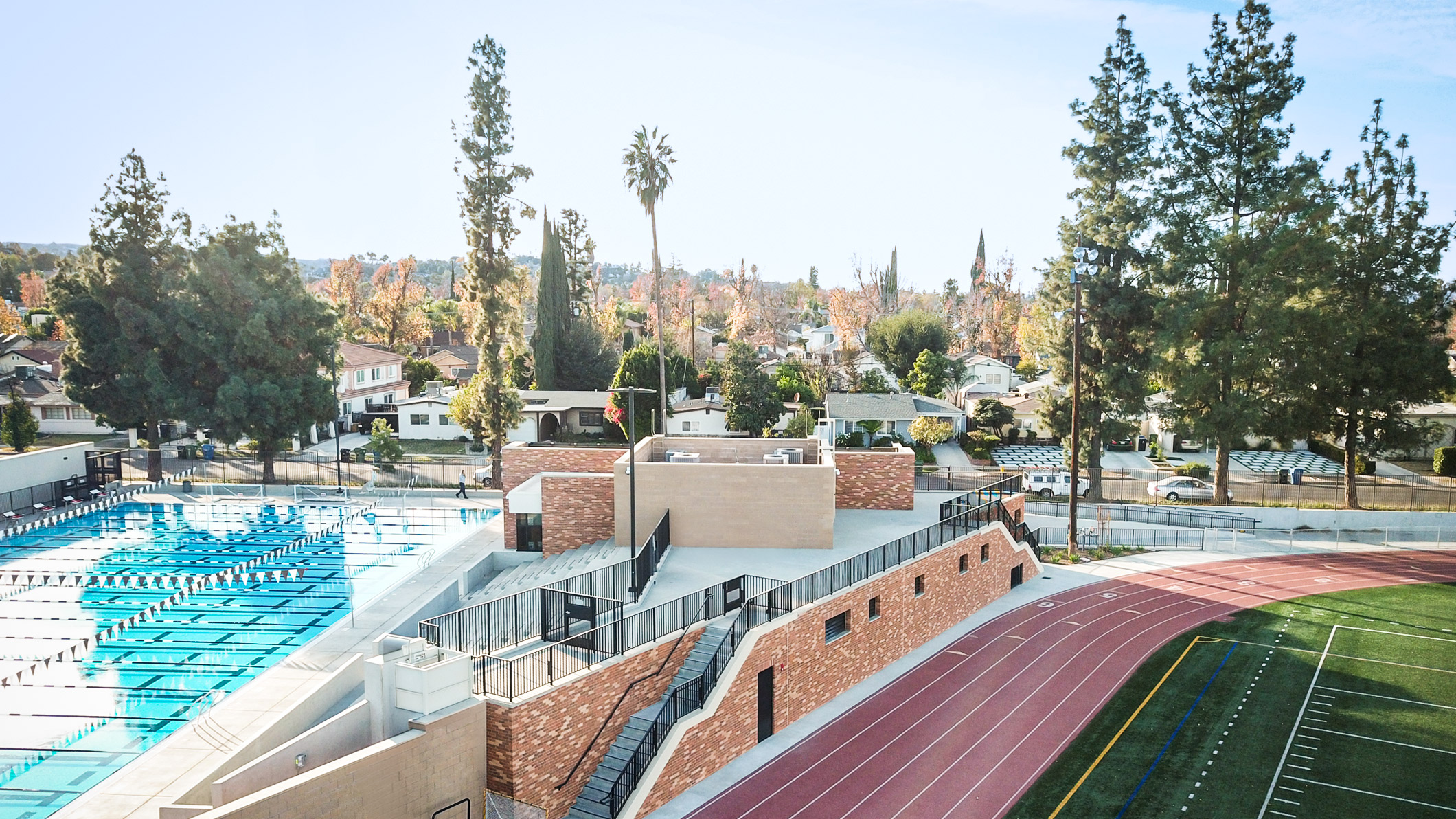 A distant, high-angle exterior of the Robinson Family Aquatics Center at Crespi Carmelite High School in Encino, a Pica+Sullivan Southern California educational architectural project. The angle showcases the Center's proximity to the track and football field.